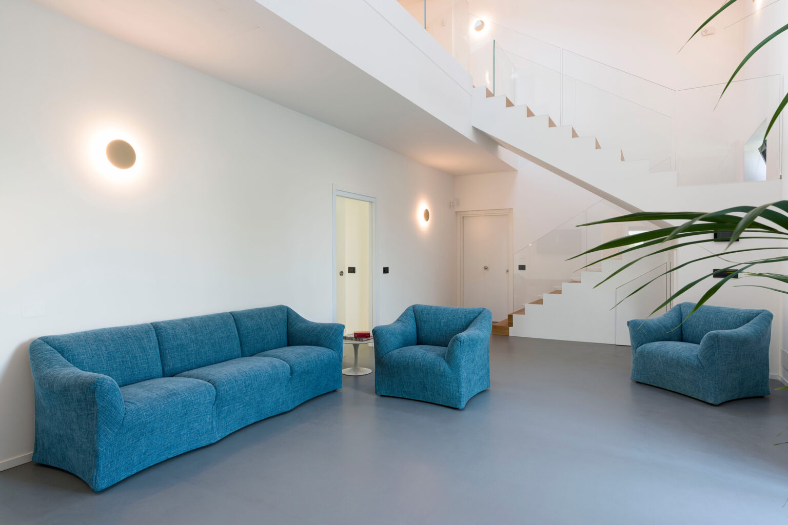 Modern living room featuring a blue sofa and armchairs, minimalist design with white walls and a staircase, natural light illuminating the space. | Only on SopranoVillas
