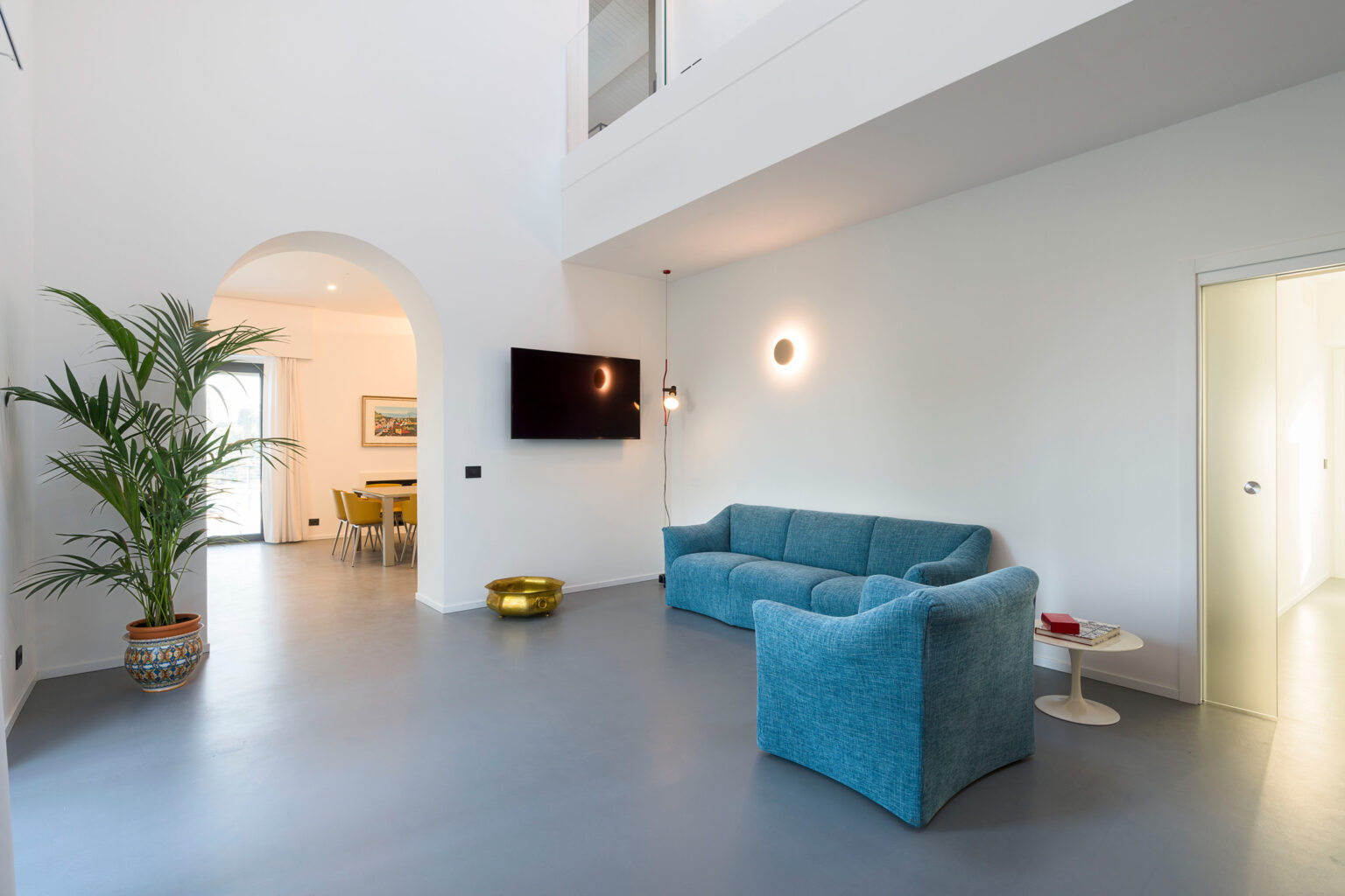 Modern living room featuring a turquoise sofa, a large potted plant, and a wall-mounted TV. The space is illuminated by stylish lighting fixtures, with an adjacent dining area visible through an archway. The sleek, gray floor complements the minimalist design and bright white walls. | Only on SopranoVillas