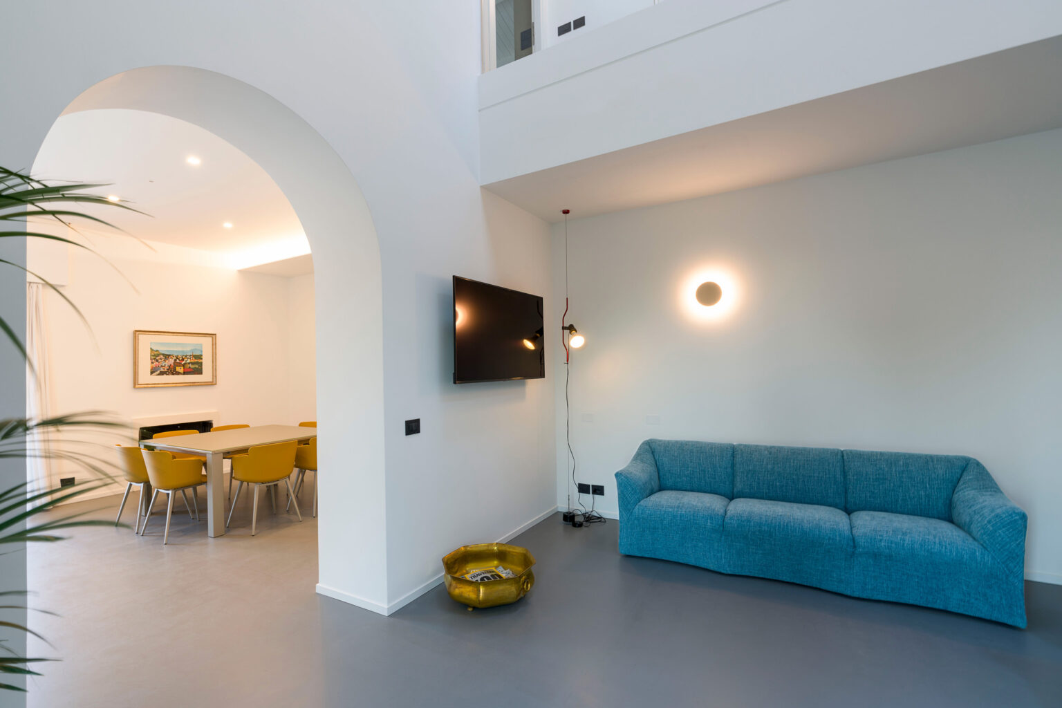 Modern living room interior featuring a light blue sofa, a wall-mounted television, and a dining area with yellow chairs. The space is bright and airy, highlighted by natural light and minimalist decor elements. | Only on SopranoVillas