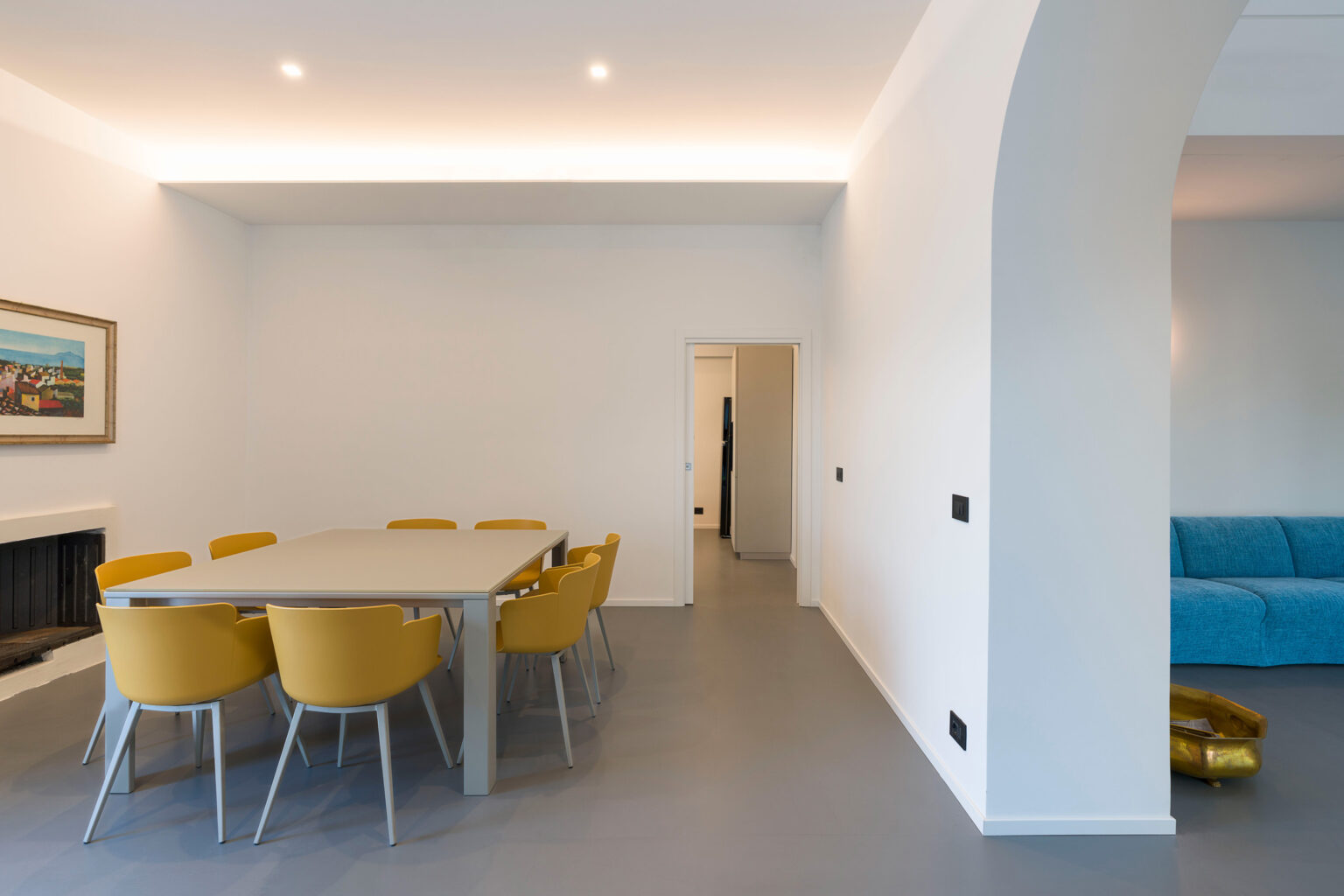 Modern dining area featuring a large table surrounded by yellow chairs, with a minimalist design and soft lighting. A blue sofa is visible in the adjacent living space, creating a bright and inviting atmosphere. | Only on SopranoVillas