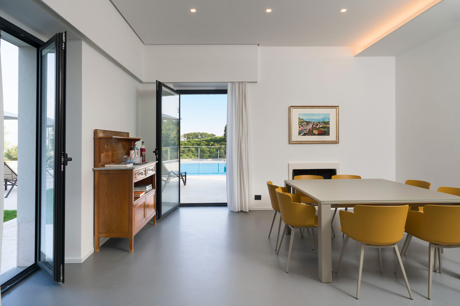Modern dining area featuring a light-filled space with a large table and yellow chairs, overlooking a pool and greenery through sliding glass doors. A wooden sideboard and framed artwork add a touch of warmth to the minimalist design. | Only on SopranoVillas