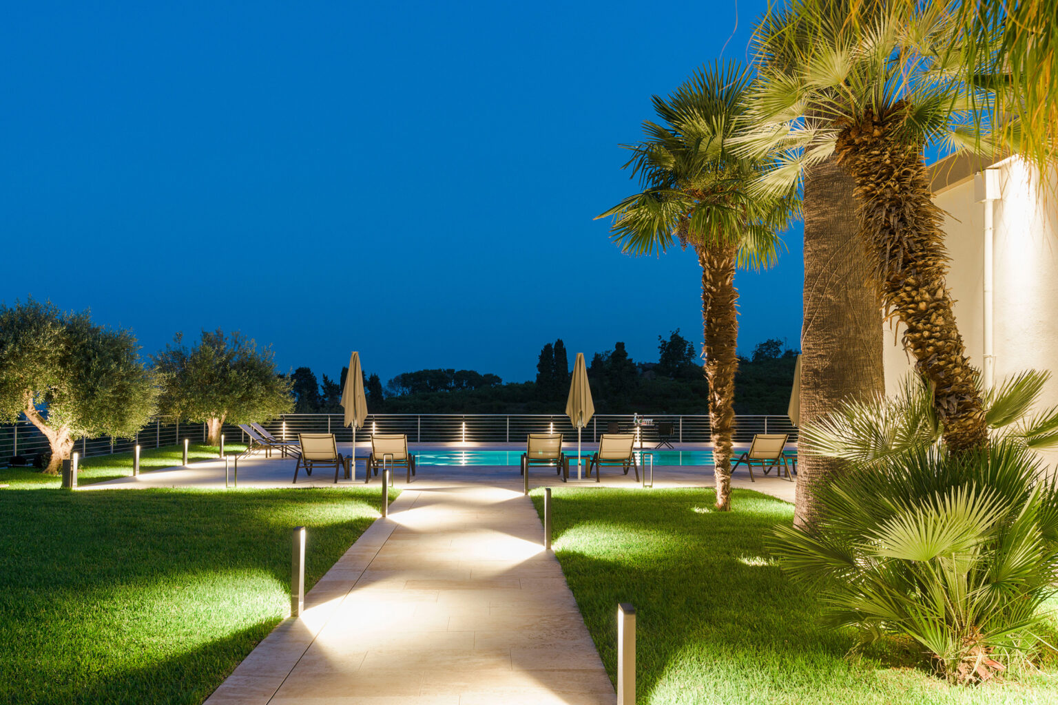 Illuminated pathway leading to a serene pool area surrounded by palm trees and olive trees at dusk, showcasing a relaxing outdoor space perfect for evening gatherings. | Only on SopranoVillas