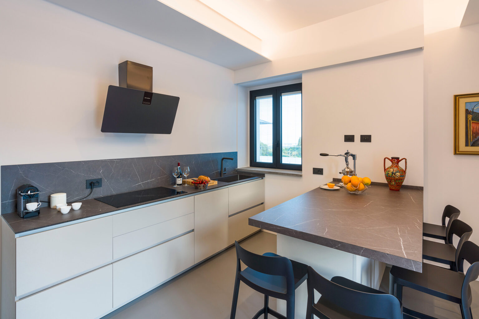 Modern kitchen interior featuring sleek cabinetry, a central island with fruit bowl, and contemporary appliances. The space is well-lit with a large window offering a view, complemented by minimalist decor and stylish seating. | Only on SopranoVillas