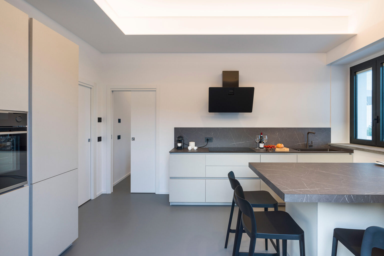 Modern kitchen interior featuring sleek white cabinetry, a dark countertop, and minimalist design. The space includes a wall-mounted television, contemporary bar stools, and large windows allowing natural light. | Only on SopranoVillas