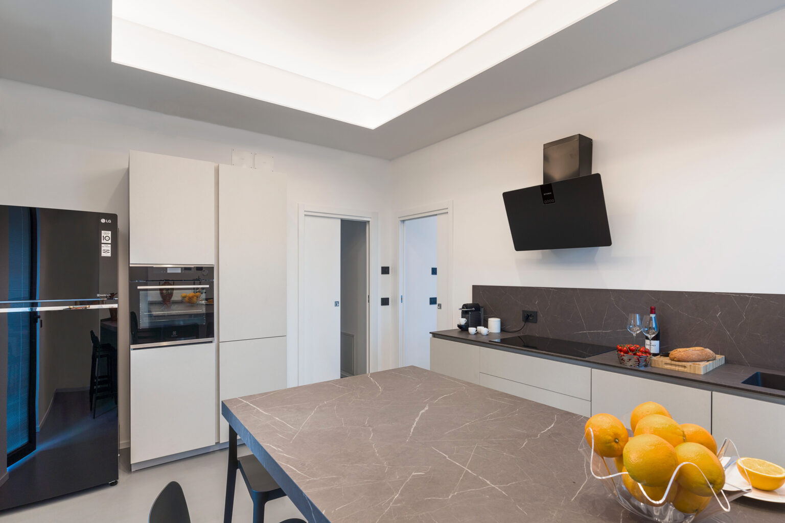 Modern kitchen featuring sleek cabinetry, a large island with a marble countertop, and a wall-mounted TV. The space includes a stainless steel refrigerator, coffee maker, and a bowl of oranges, illuminated by recessed ceiling lights. | Only on SopranoVillas