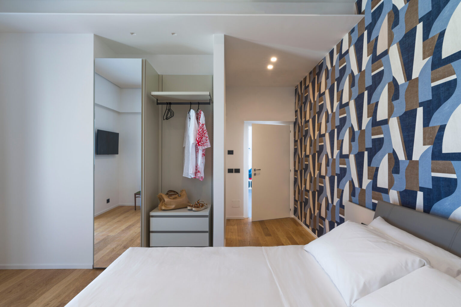 Modern bedroom featuring a stylish patterned accent wall, wooden flooring, a neatly made bed, and an open closet with clothes and accessories, creating a cozy and inviting atmosphere. | Only on SopranoVillas