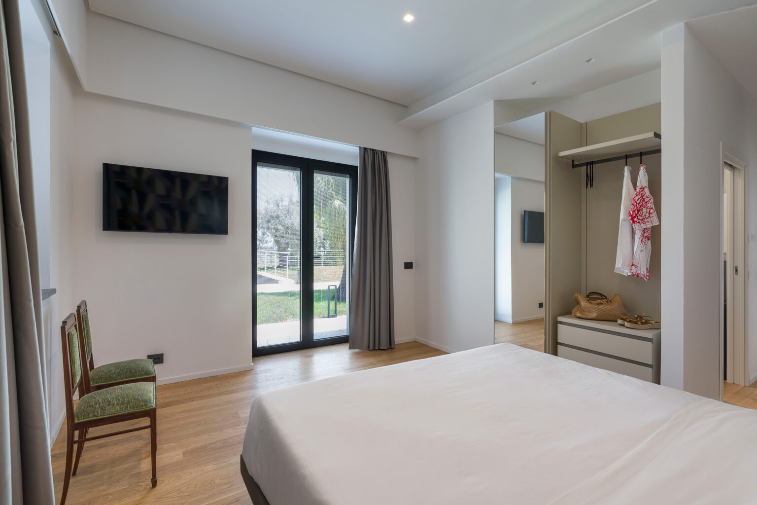 Modern bedroom interior featuring a double bed, wooden flooring, a wall-mounted TV, and large glass doors leading to an outdoor area. The room includes a chair, a mirror closet with hangers, and stylish decor elements, creating a bright and inviting atmosphere. | Only on SopranoVillas