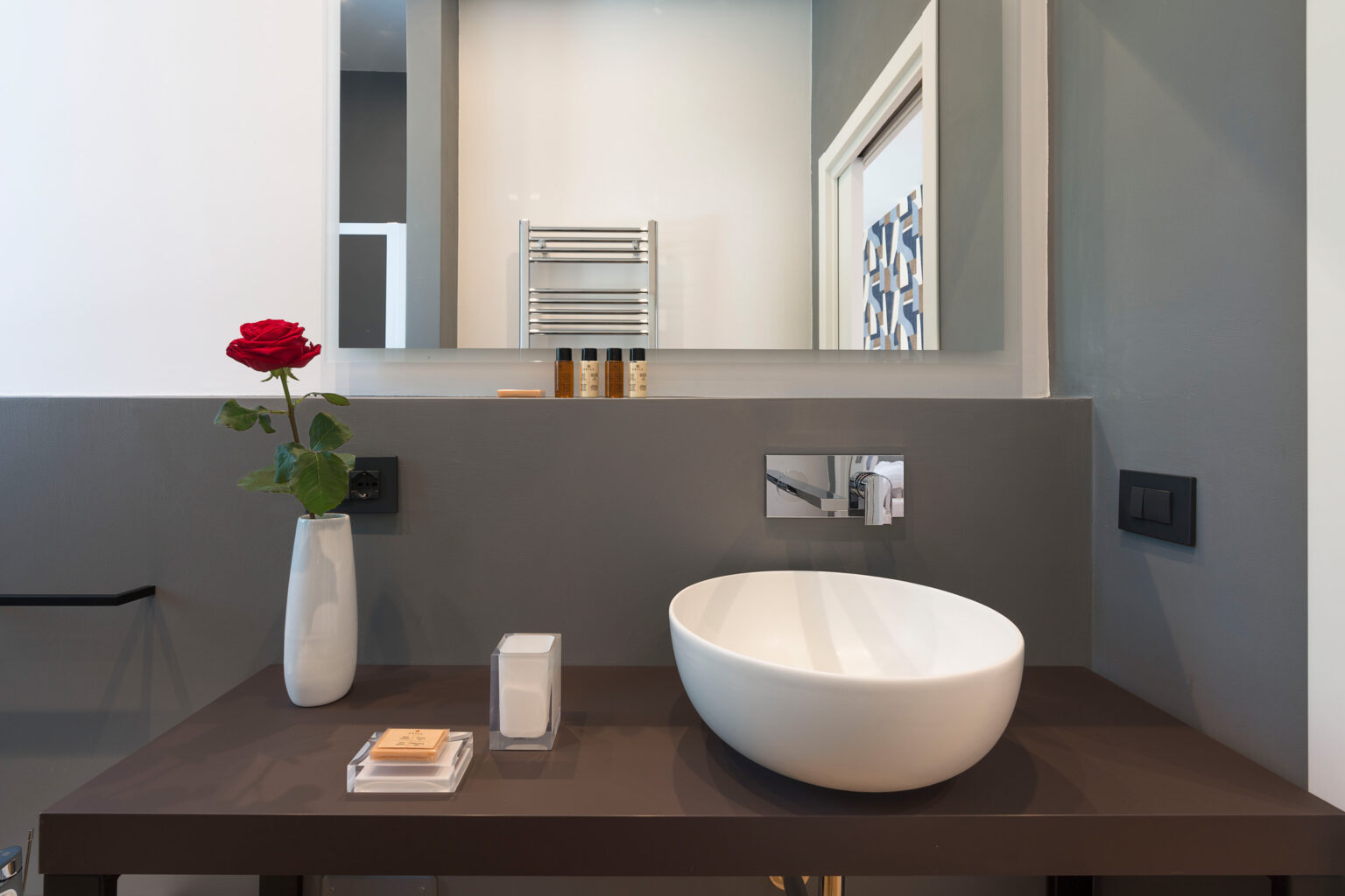 Modern bathroom interior featuring a minimalist design with a white vessel sink, sleek brown countertop, and a decorative red rose in a vase. The space includes a wall-mounted mirror, stylish faucet, and neatly arranged toiletries, creating a chic and serene atmosphere. | Only on SopranoVillas