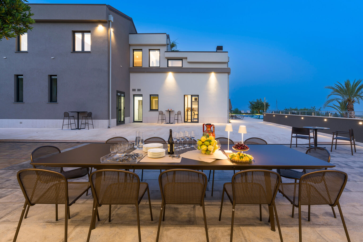 Outdoor dining area with a large table set for a meal, surrounded by modern chairs, against a backdrop of a contemporary home at dusk. The scene features elegant table settings with fruit and wine, highlighting a stylish and inviting atmosphere. | Only on SopranoVillas