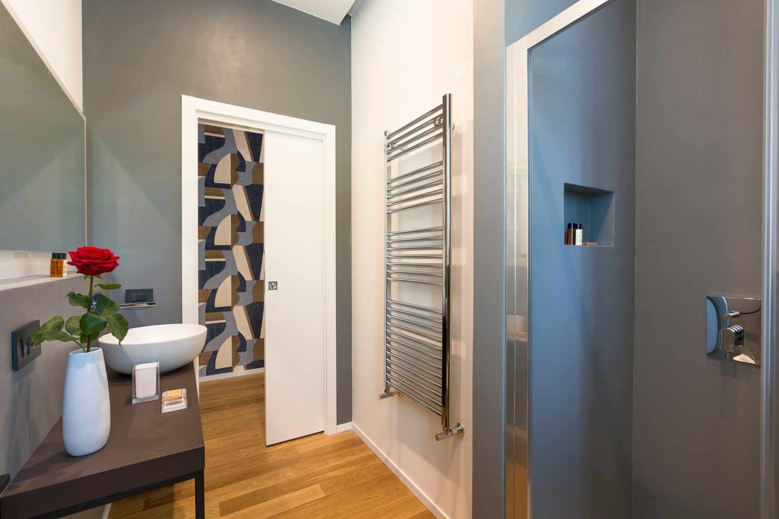 Modern bathroom interior featuring a sleek design with a white vessel sink, a decorative red rose, a towel warmer, and a glass shower. The walls are painted in a combination of gray and blue tones, complemented by a stylish patterned door. Natural wood flooring adds warmth to the space. | Only on SopranoVillas