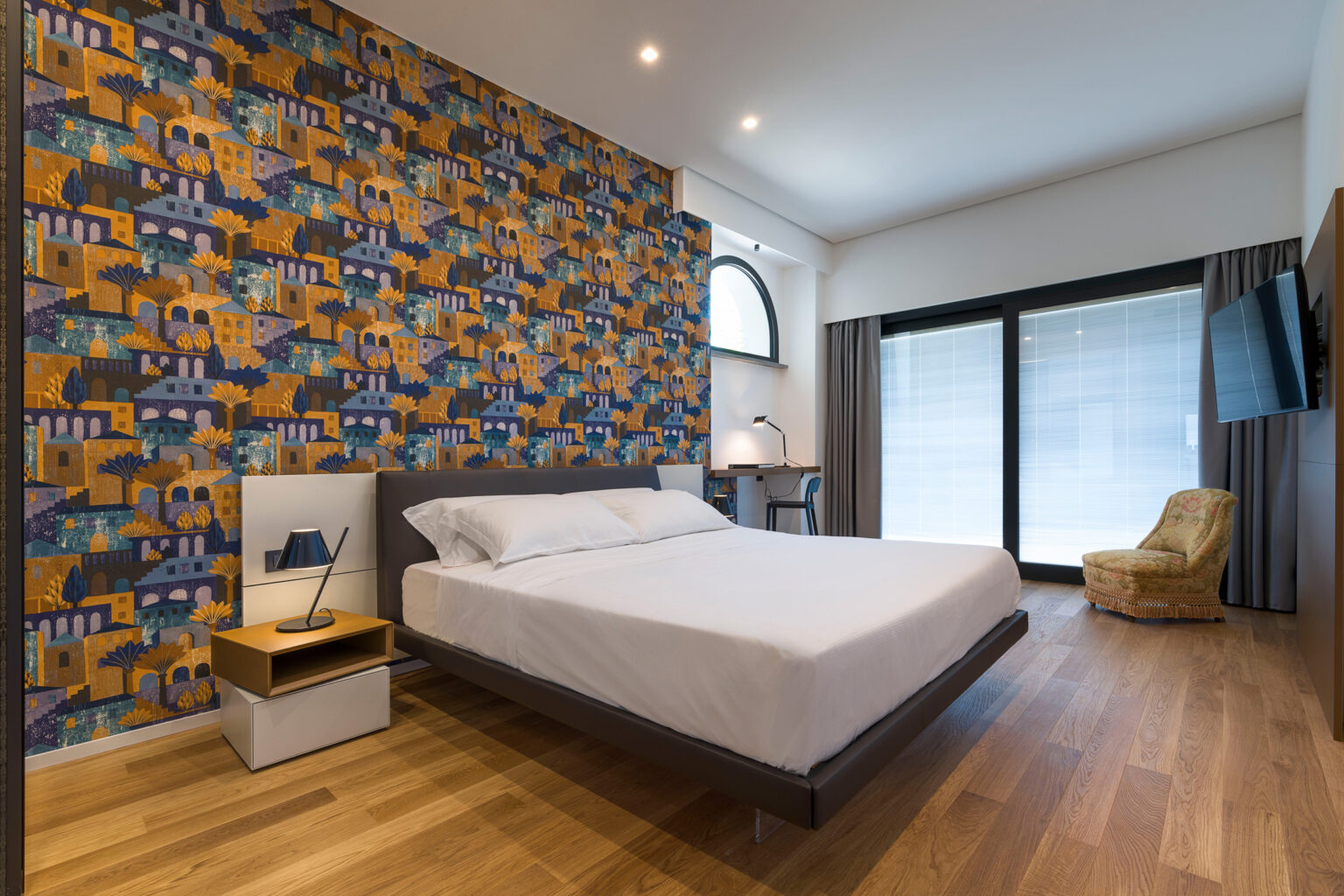 Modern bedroom featuring a stylish bed with white linens, a decorative patterned accent wall in warm colors, a bedside lamp, a small desk with a chair, and a cozy armchair. Large windows allow natural light, enhancing the contemporary design and inviting atmosphere. | Only on SopranoVillas