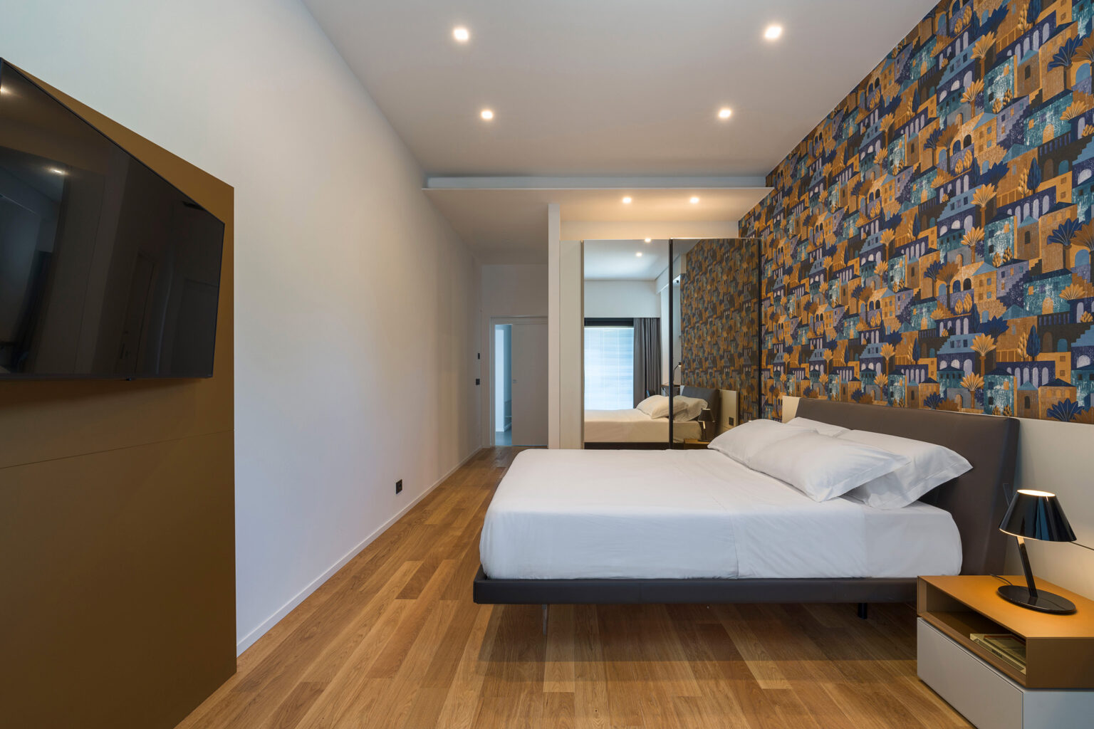 Modern bedroom featuring a minimalist design with a large bed, stylish lamp, and a mounted flat-screen TV. The accent wall displays a vibrant patterned wallpaper, while natural light filters through a nearby window, enhancing the warm wooden flooring. | Only on SopranoVillas