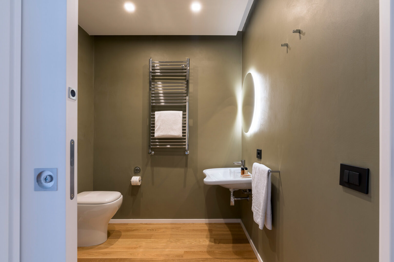 Modern bathroom featuring a toilet, towel rack, and minimalist design with green walls and wooden flooring, illuminated by stylish lighting. | Only on SopranoVillas