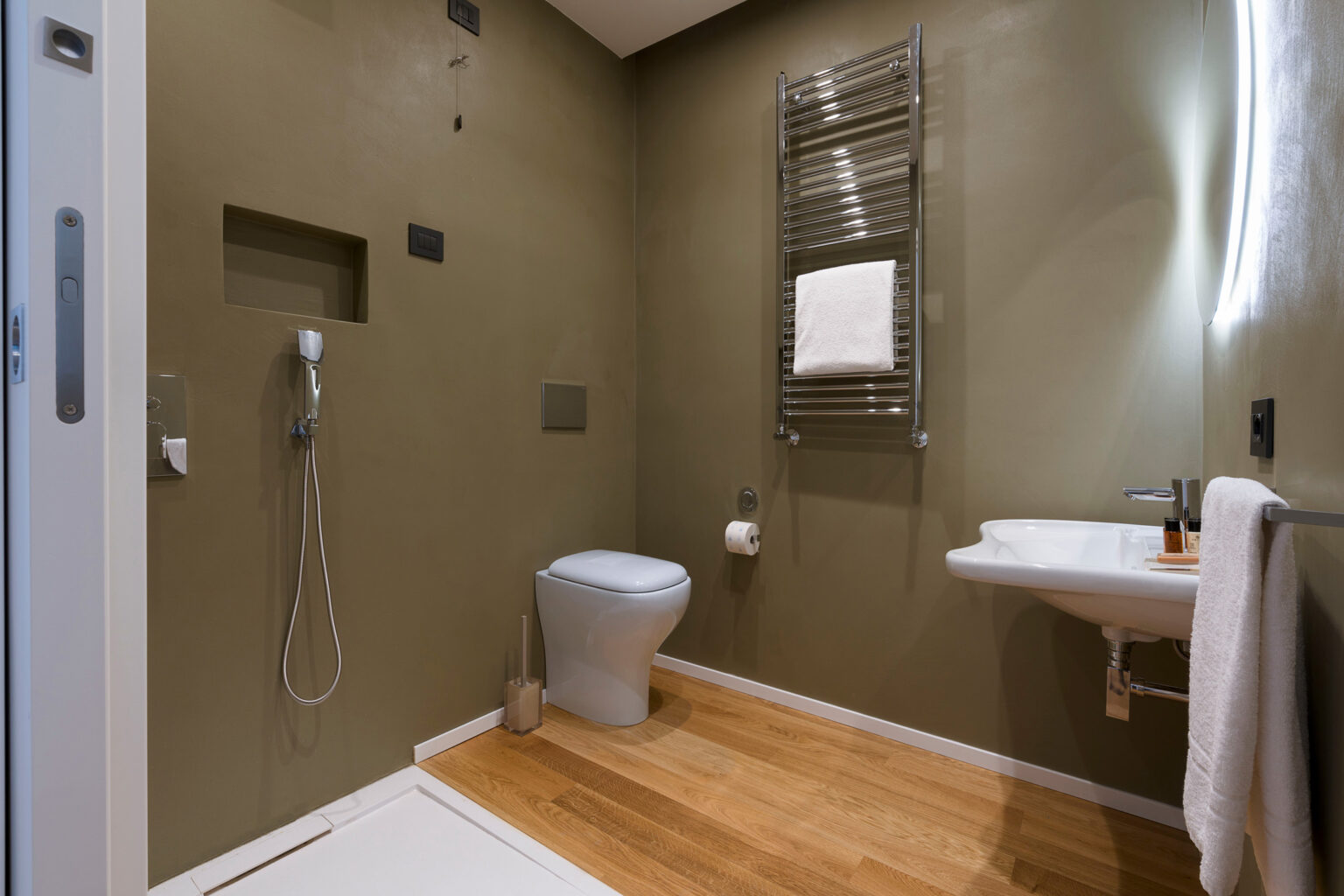 Modern bathroom design featuring green walls, a sleek toilet, a wall-mounted sink, and a towel radiator, complemented by wooden flooring for a warm aesthetic. | Only on SopranoVillas