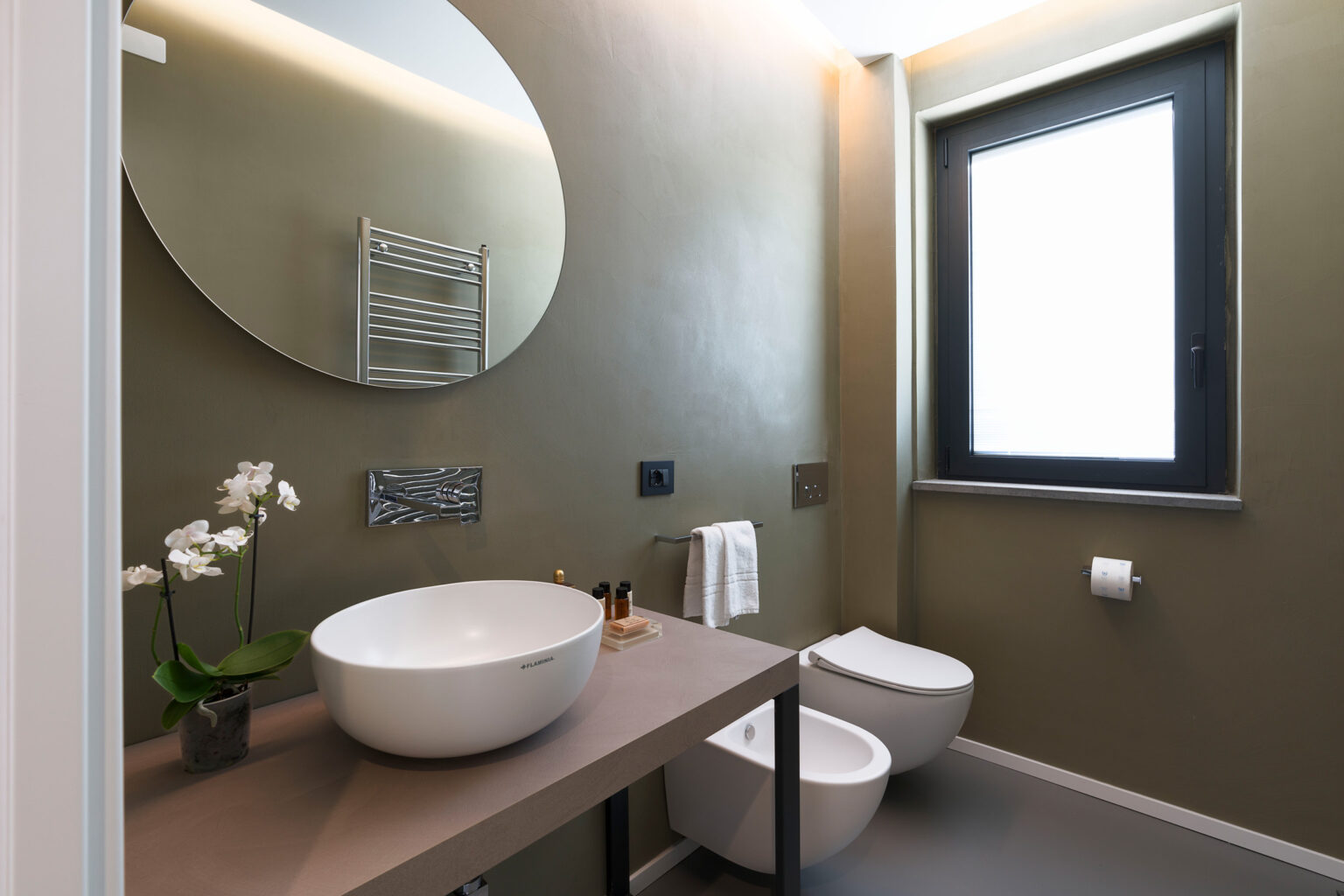 Modern bathroom featuring a round mirror, minimalist sink, and stylish fixtures. The space is adorned with a potted orchid and has a neutral color palette, creating a serene atmosphere. Natural light streams in through a window. | Only on SopranoVillas