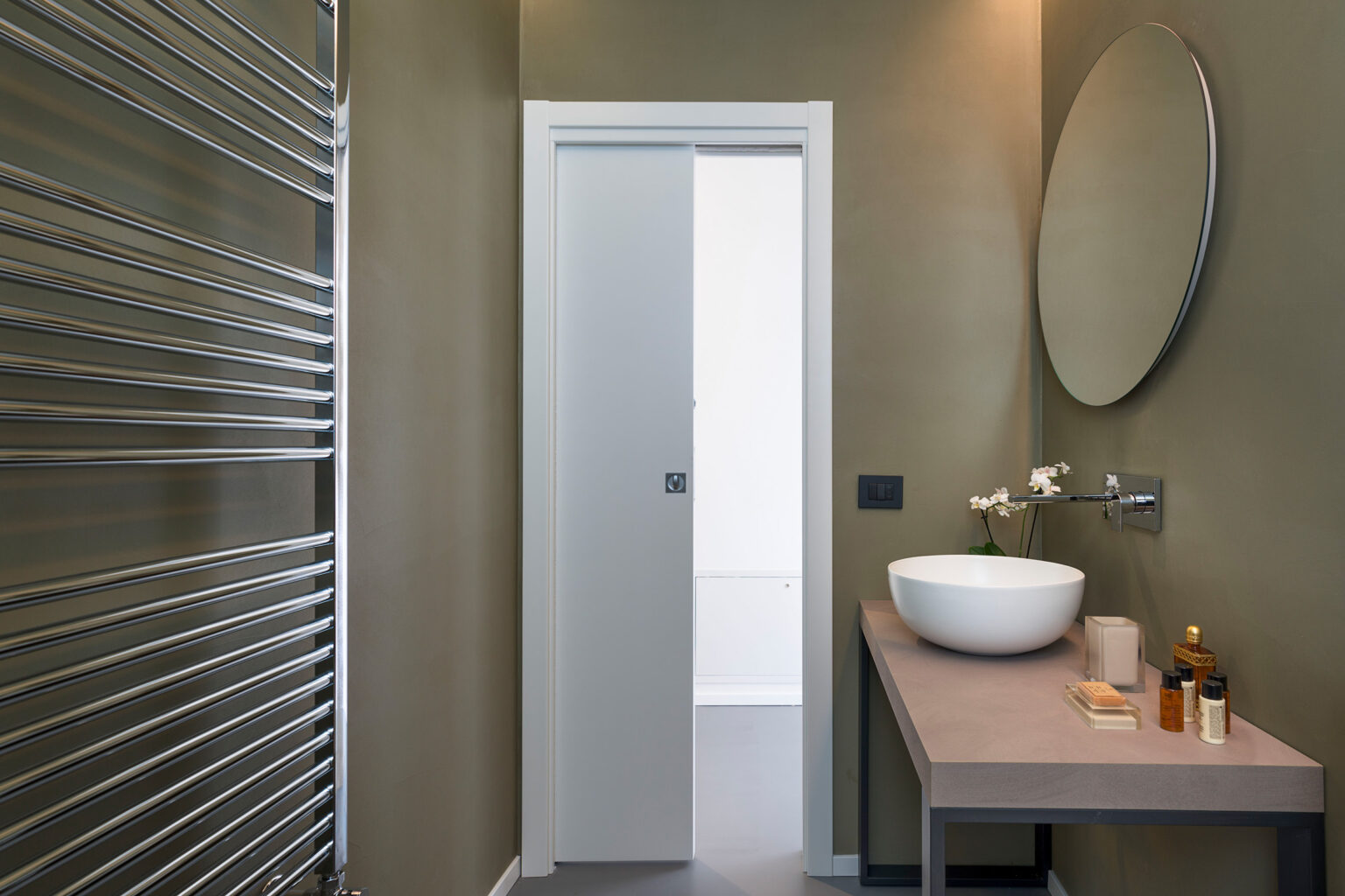 Modern bathroom design featuring a sleek sliding door, minimalist round mirror, and a stylish countertop with a white vessel sink. The walls are painted in a muted green tone, complemented by a contemporary towel radiator and elegant decor items. | Only on SopranoVillas