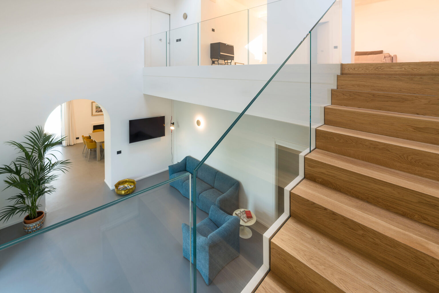 Modern interior design featuring a spacious living area with a blue sofa, wooden staircase, and glass railing. Natural light floods the space, highlighting the minimalist decor and indoor plants. Ideal for showcasing contemporary home aesthetics. | Only on SopranoVillas