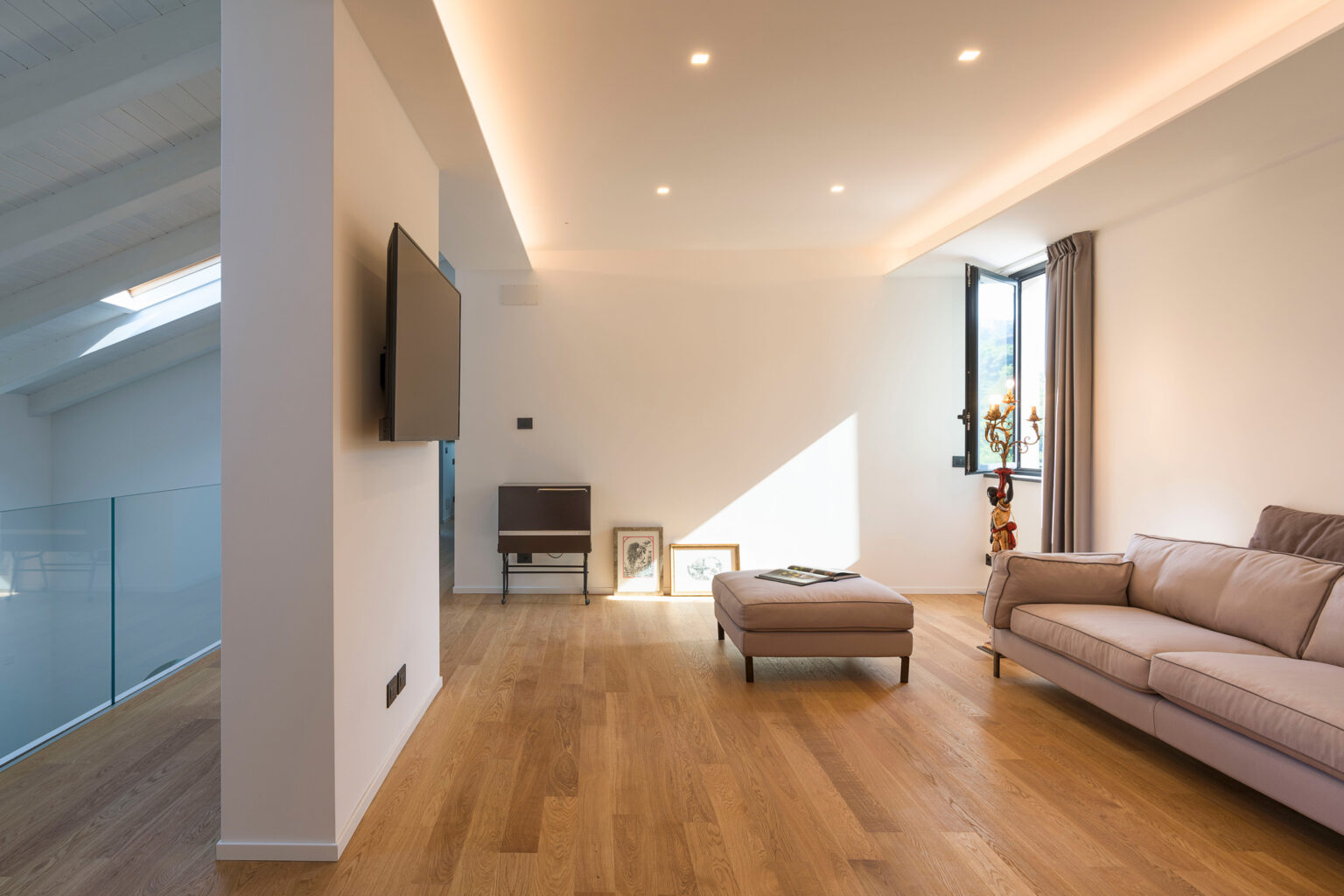 Modern living room with natural light, featuring a beige sofa, wooden flooring, and minimalist decor. Wall-mounted TV and large window enhance the spacious feel. | Only on SopranoVillas
