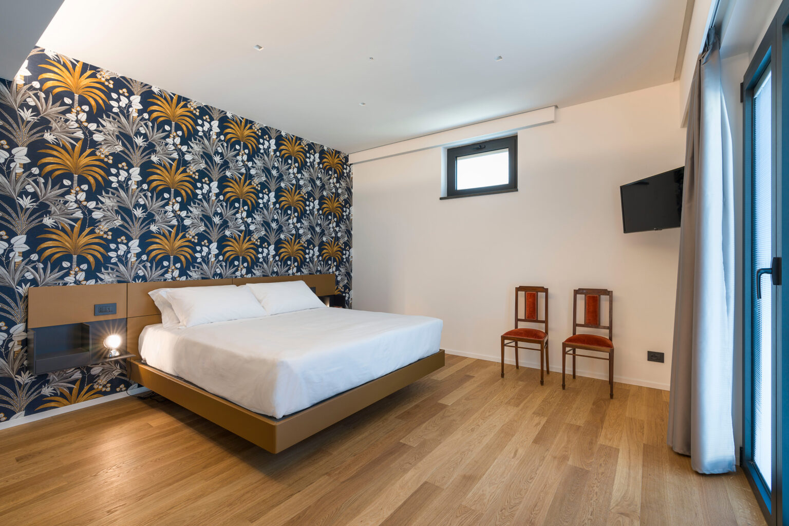 Modern hotel room featuring a double bed, stylish floral wallpaper, hardwood flooring, and two wooden chairs, with a flat-screen TV mounted on the wall. Natural light streams in through a window, creating a welcoming atmosphere. | Only on SopranoVillas