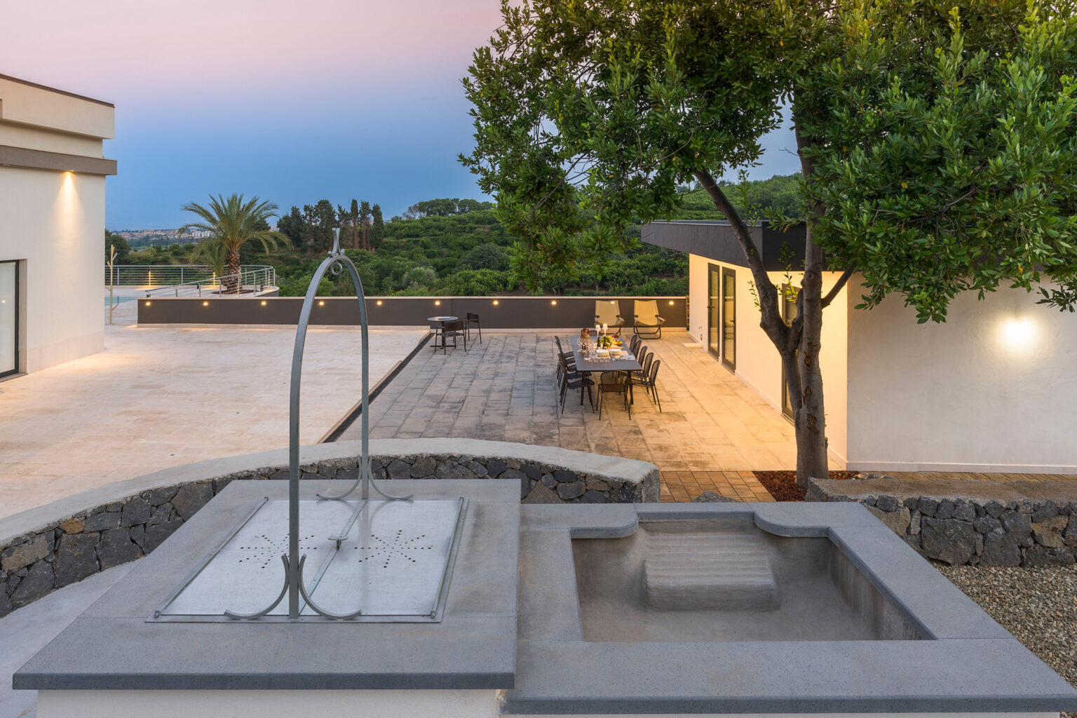Modern outdoor living space featuring a sleek water fountain, dining area, and lush greenery, set against a serene twilight backdrop. Ideal for relaxation and entertaining. | Only on SopranoVillas