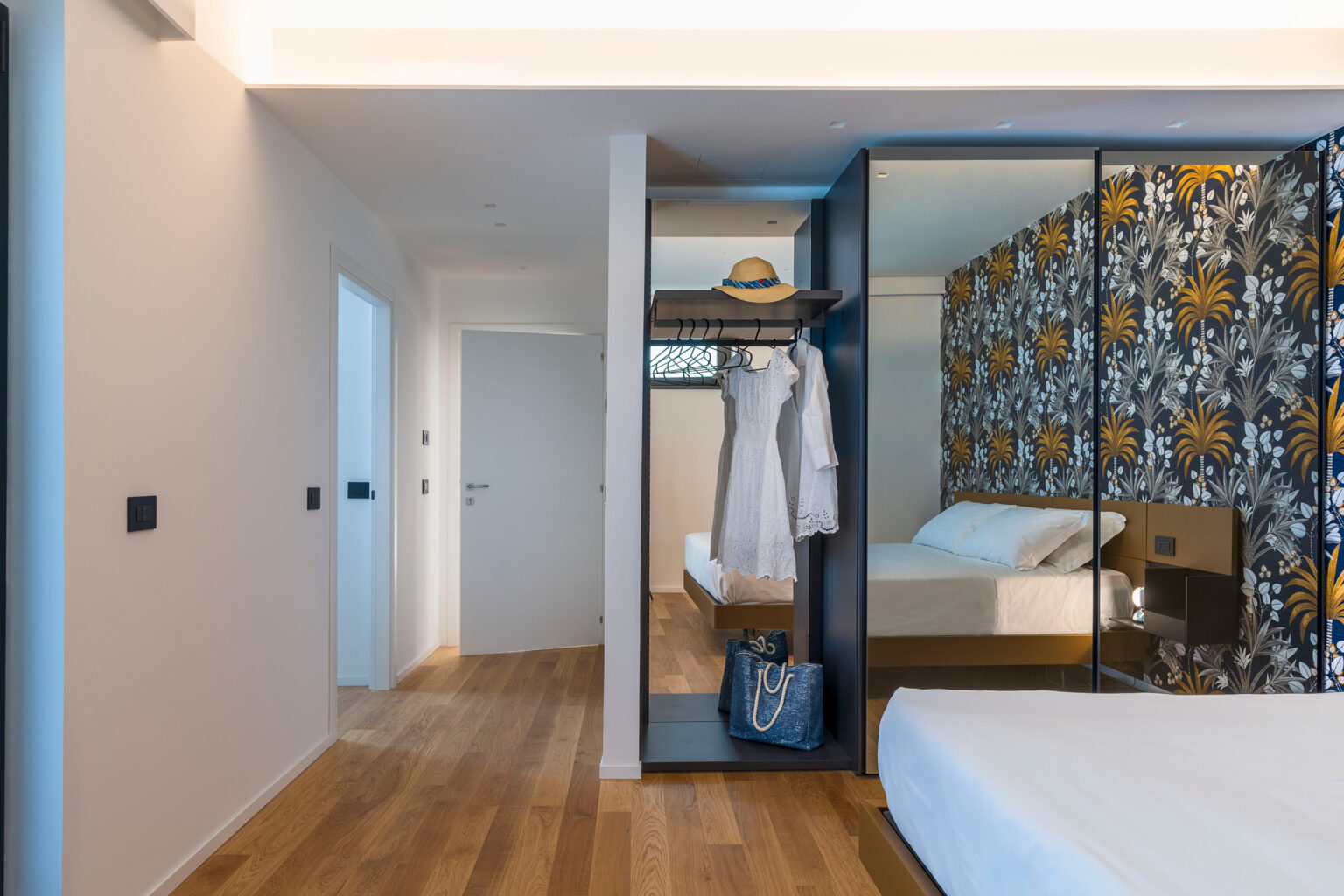 Modern bedroom interior featuring a stylish closet with a white dress and a straw hat, complemented by floral wallpaper and wooden flooring. The room includes a comfortable bed, creating a cozy and elegant atmosphere. | Only on SopranoVillas