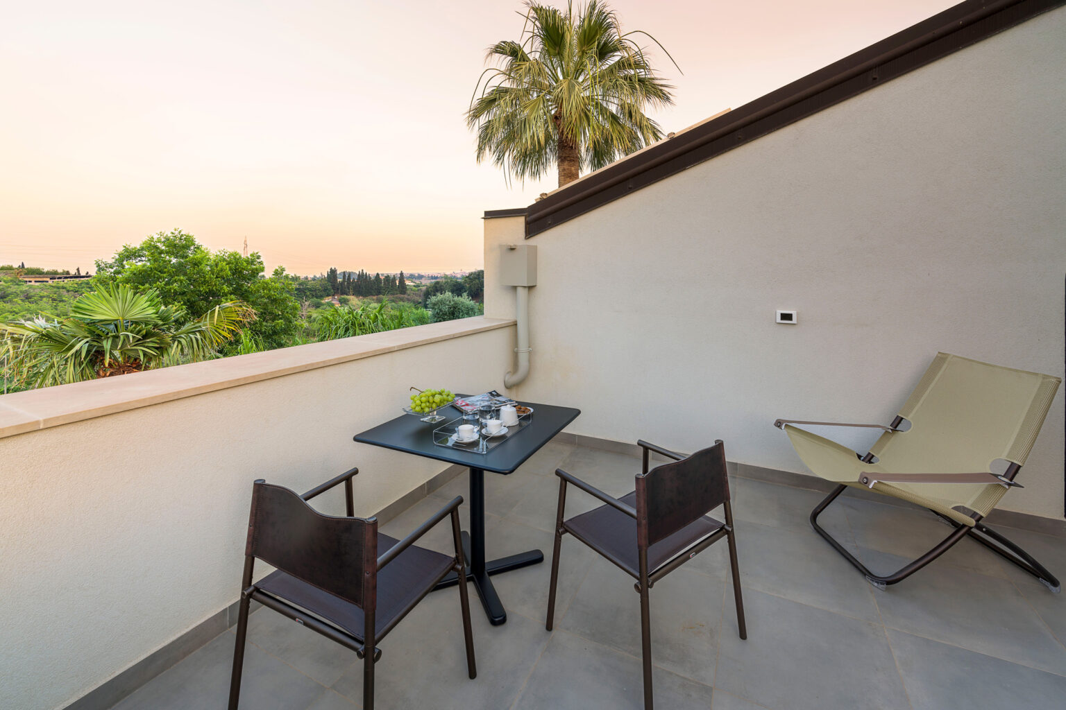 Cozy balcony with a small table set for two, featuring a coffee service and fresh grapes, surrounded by lush greenery and a palm tree, against a serene sunset backdrop. Ideal for relaxation and enjoying outdoor views. | Only on SopranoVillas