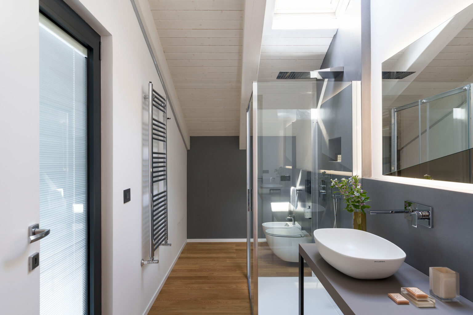 Modern bathroom interior featuring a sleek glass shower, a freestanding oval sink, and wooden flooring. The design includes a towel radiator, minimalist decor, and natural light from a skylight, creating a bright and airy atmosphere. | Only on SopranoVillas