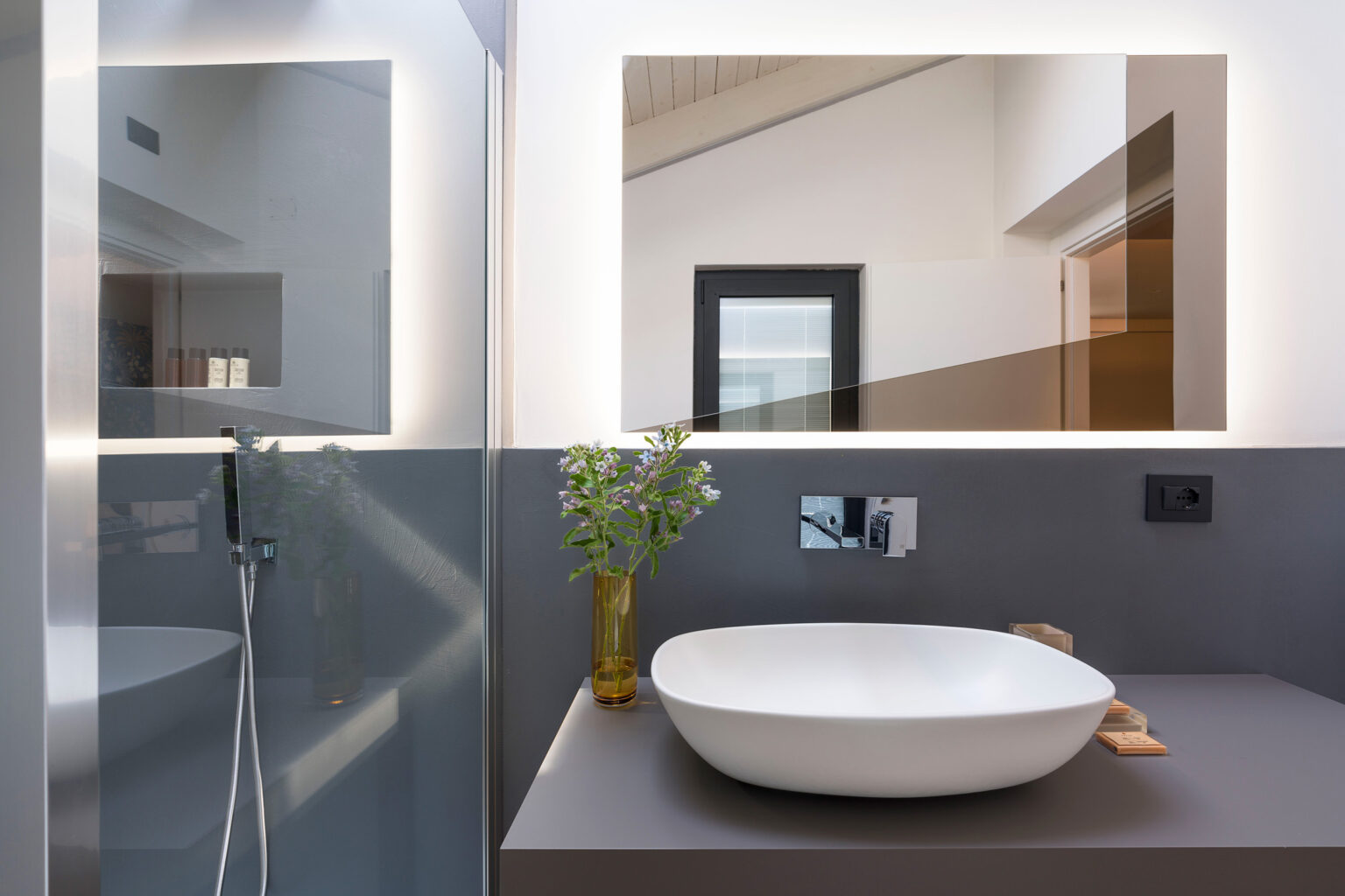 Modern bathroom interior featuring a sleek white vessel sink, stylish faucet, and a minimalist design. The space includes a contemporary mirror with integrated lighting, a shower area with glass partition, and a decorative vase with greenery, creating a fresh and inviting atmosphere. | Only on SopranoVillas