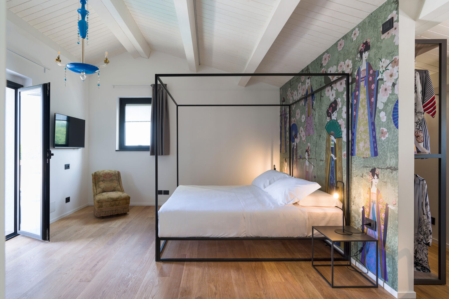Modern bedroom featuring a stylish four-poster bed, hardwood flooring, and vibrant wall art. The room includes a cozy chair, a television, and large windows for natural light. Ideal for a relaxing stay. | Only on SopranoVillas
