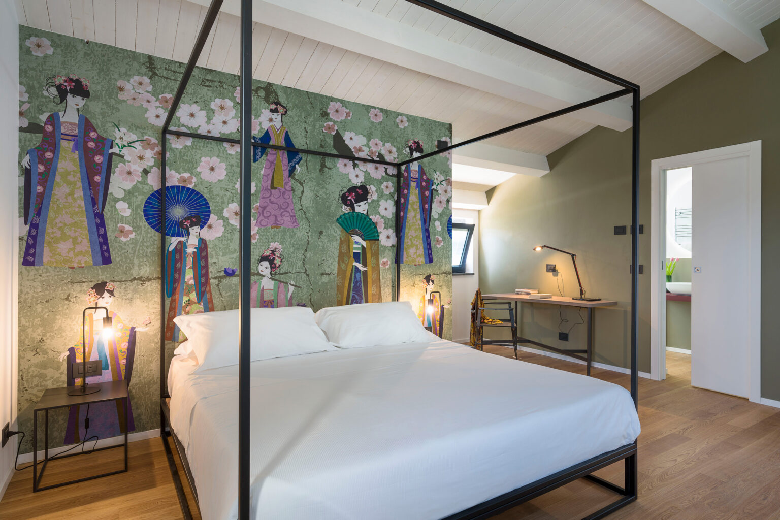 Modern bedroom featuring a four-poster bed with a minimalist design, adorned with a vibrant floral mural depicting traditional figures. The room has wooden flooring, stylish bedside lamps, and a workspace with a desk, creating a cozy and artistic atmosphere. | Only on SopranoVillas