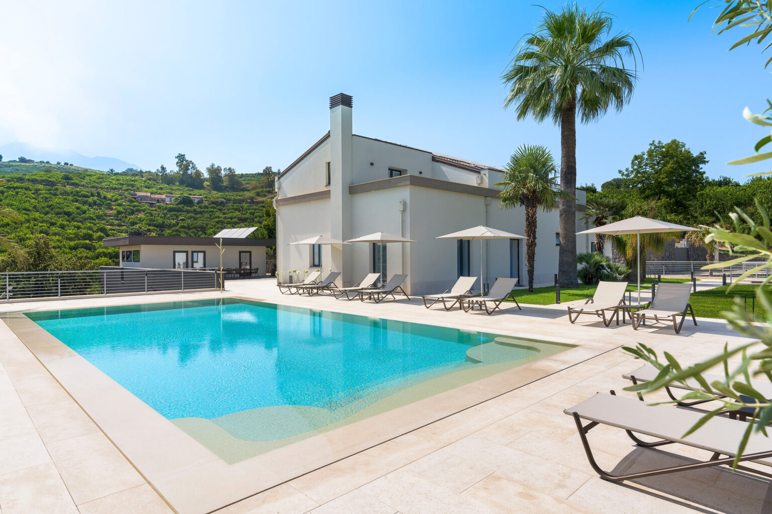 Modern villa with a swimming pool surrounded by lounge chairs and umbrellas, set against a backdrop of lush greenery and blue skies. Ideal for luxury vacation rentals or outdoor relaxation. | Only on SopranoVillas