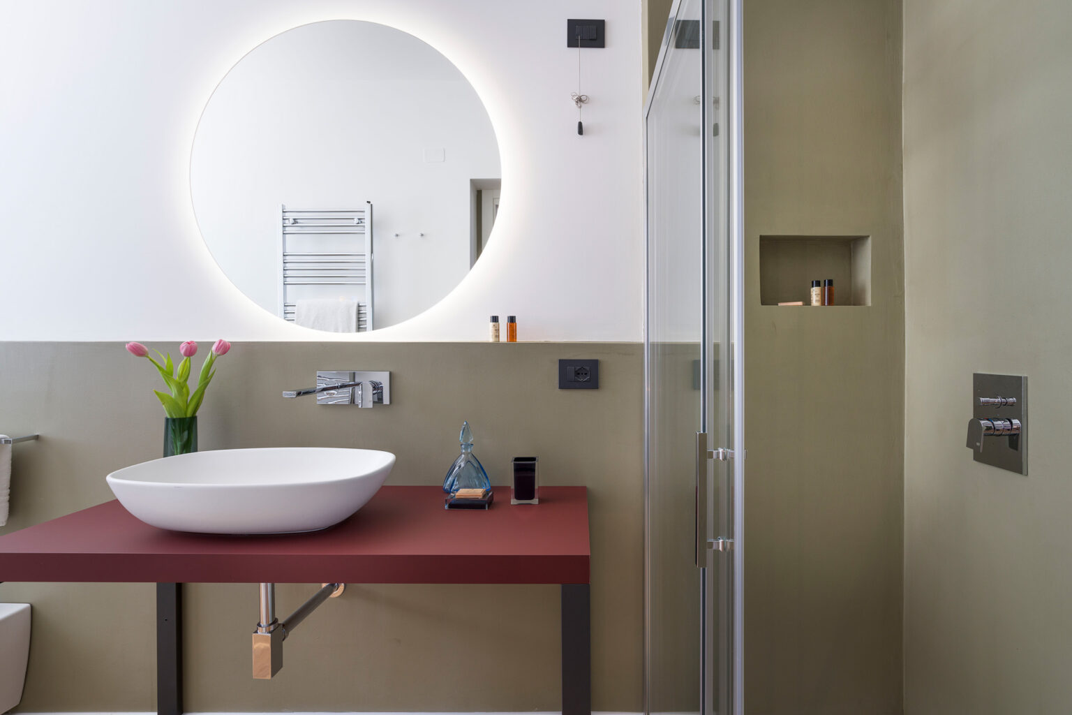 Modern bathroom interior featuring a round mirror, a stylish vessel sink on a red countertop, and a glass shower enclosure. The design includes minimalist decor with a touch of greenery and contemporary fixtures, showcasing a blend of functionality and aesthetics. | Only on SopranoVillas