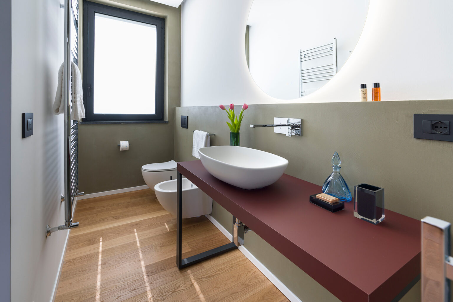 Modern bathroom design featuring a stylish wall-mounted sink on a sleek red countertop, a toilet, and a large circular mirror. Natural light flows in through a window, complemented by minimalist decor and green accent walls, creating a fresh and inviting atmosphere. | Only on SopranoVillas