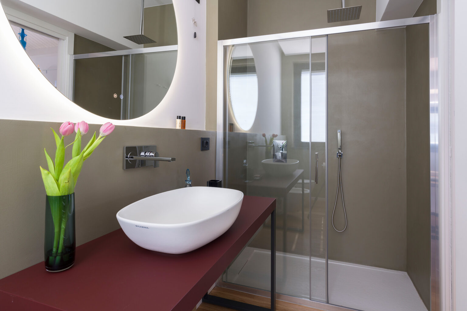 Modern bathroom interior featuring a stylish vessel sink on a burgundy countertop, a glass shower enclosure, and a round mirror. Fresh flowers in a vase add a touch of elegance to the space. | Only on SopranoVillas