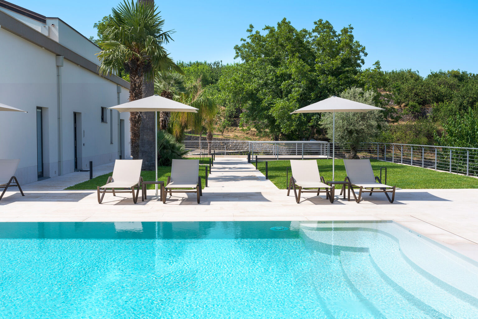 Modern outdoor swimming pool with lounge chairs and umbrellas, surrounded by lush greenery and a stylish patio area. Ideal for relaxation and summer enjoyment. | Only on SopranoVillas