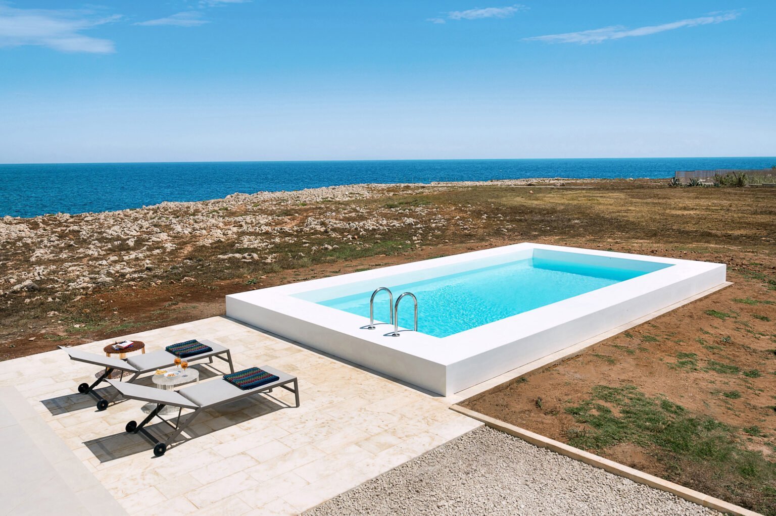 Modern outdoor swimming pool overlooking the ocean, surrounded by a grassy area and featuring lounge chairs for relaxation. Bright blue sky enhances the serene coastal setting. | Only on SopranoVillas