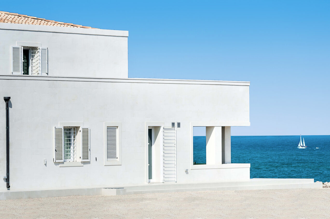 Modern coastal house with white exterior and large windows, overlooking the ocean and a sailboat in the distance, under a clear blue sky. | Only on SopranoVillas