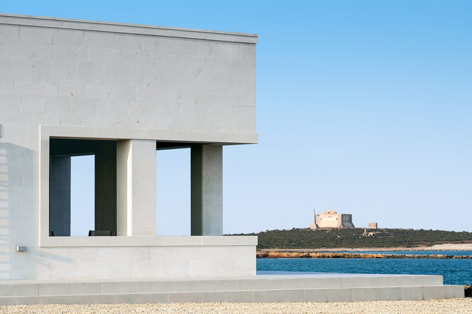 Modern architectural structure with clean lines and large windows, overlooking a serene waterfront. In the background, a historic building is visible on a distant island, set against a clear blue sky. | Only on SopranoVillas