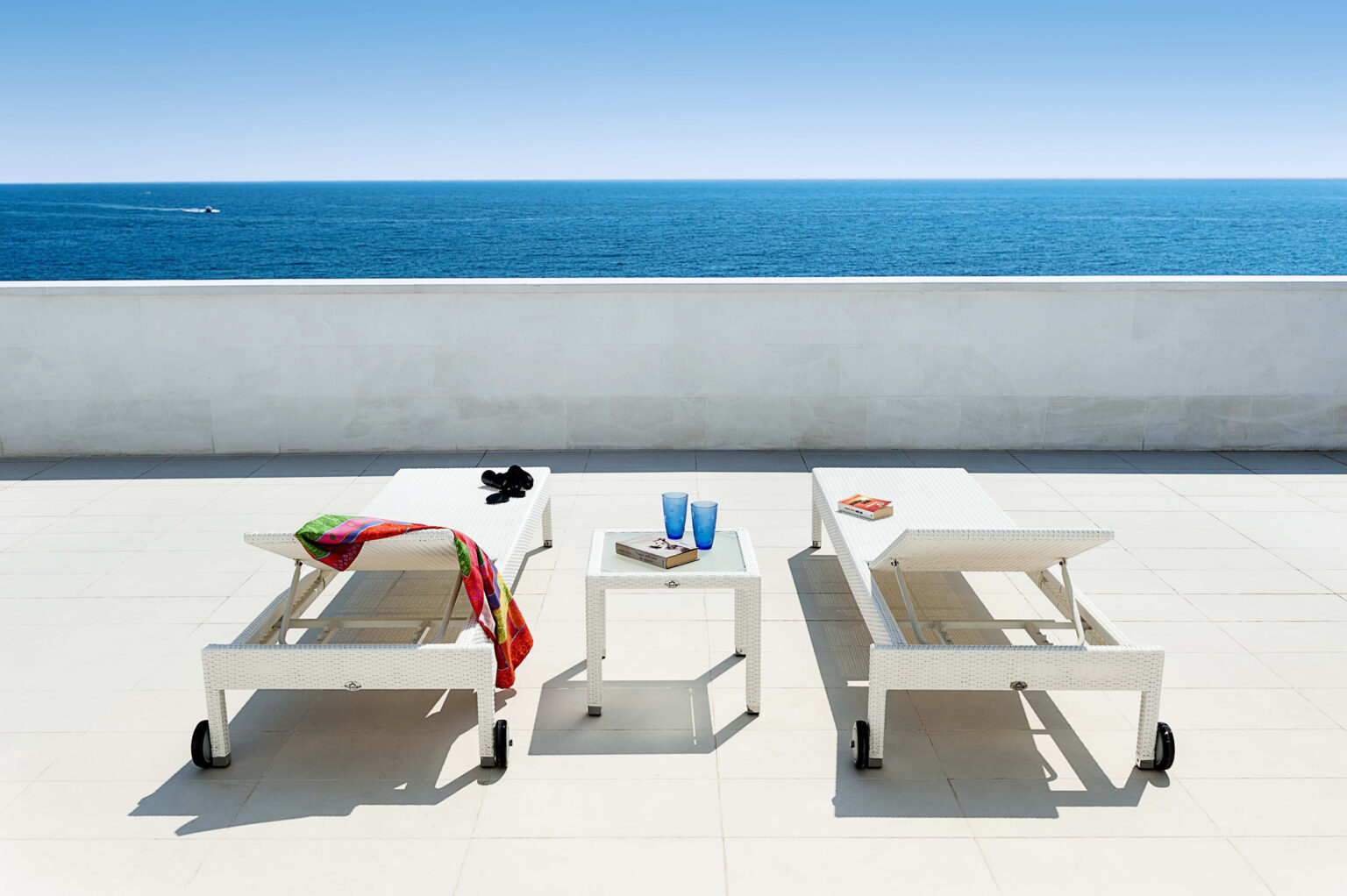 Two white lounge chairs with a colorful towel draped over one, positioned on a sunlit terrace overlooking a calm blue ocean. A small table between the chairs holds two blue glasses and a book, creating a serene coastal relaxation setting. | Only on SopranoVillas