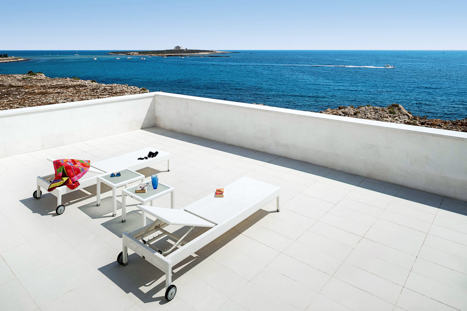 Modern outdoor terrace overlooking the blue sea, featuring white lounge chairs, a colorful beach towel, and a small table with a drink and book. Ideal for relaxation and enjoying coastal views. | Only on SopranoVillas