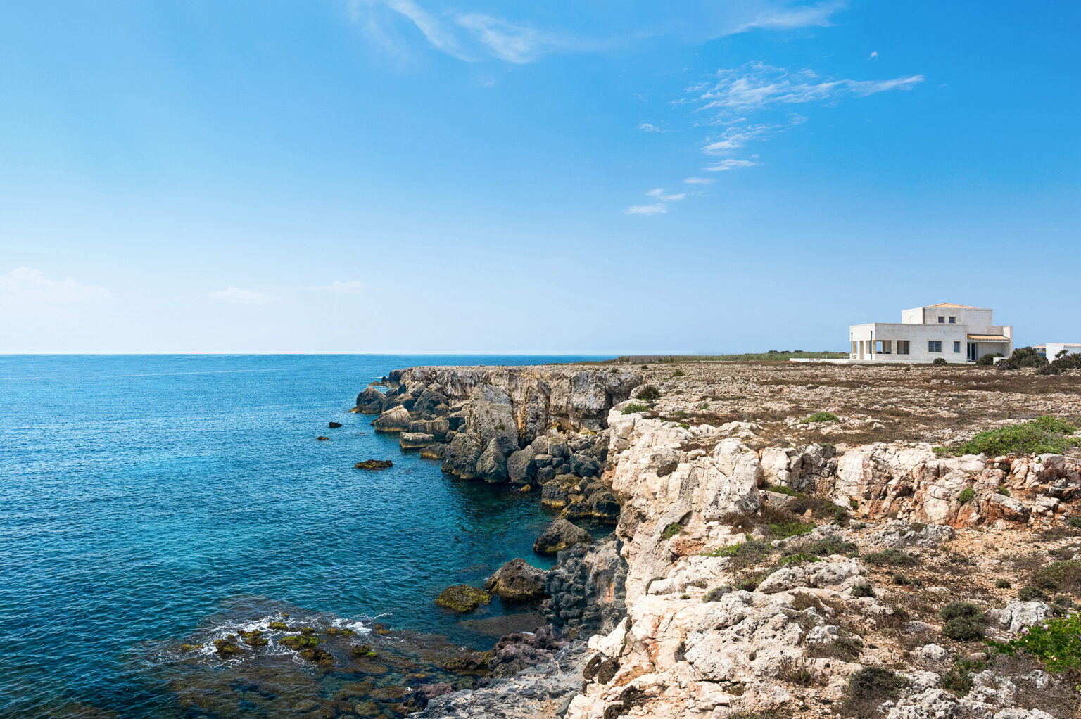 Coastal view featuring rocky cliffs and a serene blue sea under a clear sky, with a modern white house perched on the edge of the cliff, showcasing natural beauty and tranquility. | Only on SopranoVillas