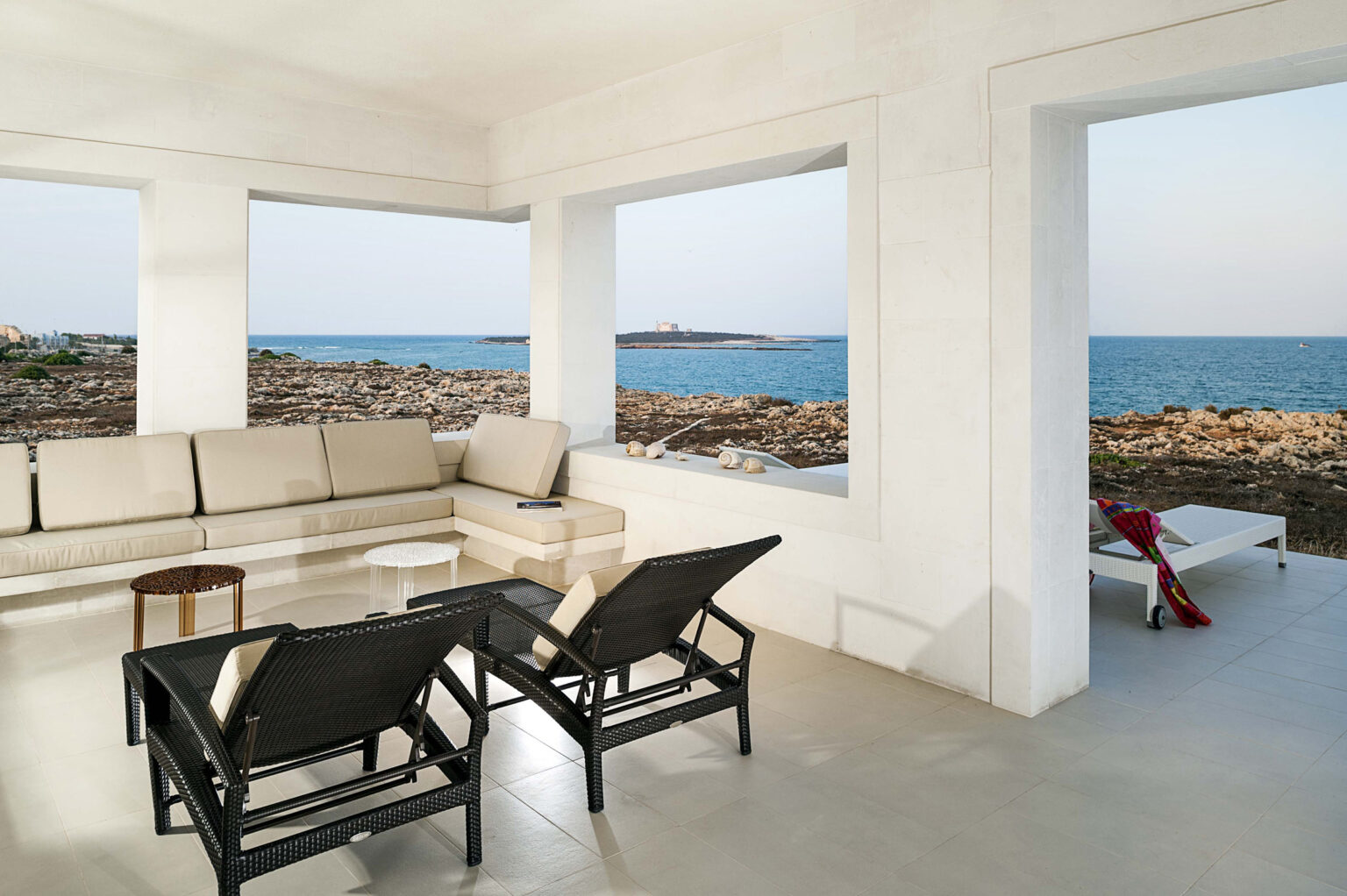 Modern coastal living space with a beige sectional sofa, black lounge chairs, and a scenic view of the ocean and rocky shoreline through large windows. The minimalist design features light-colored walls and a serene atmosphere, perfect for relaxation. | Only on SopranoVillas