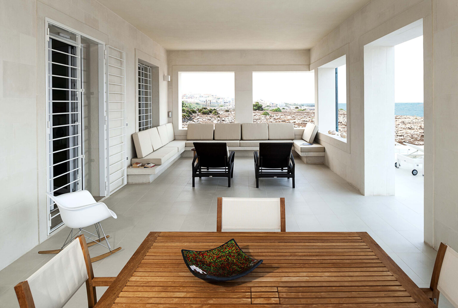 Cozy outdoor living space featuring modern furniture, including a wooden dining table and comfortable seating. The area is illuminated by natural light, with large windows offering views of the sea and rocky landscape. Ideal for relaxation and entertaining in a serene coastal setting. | Only on SopranoVillas