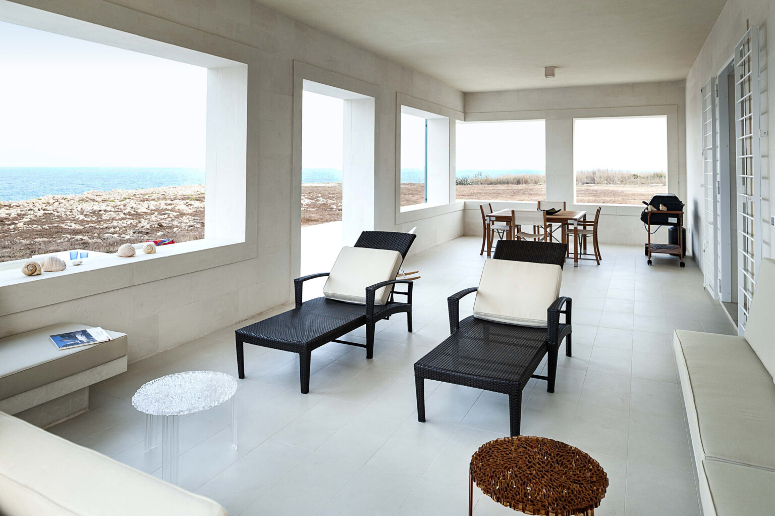 Modern coastal living room featuring minimalist design, stylish black lounge chairs, and a dining area with ocean views through large windows. | Only on SopranoVillas