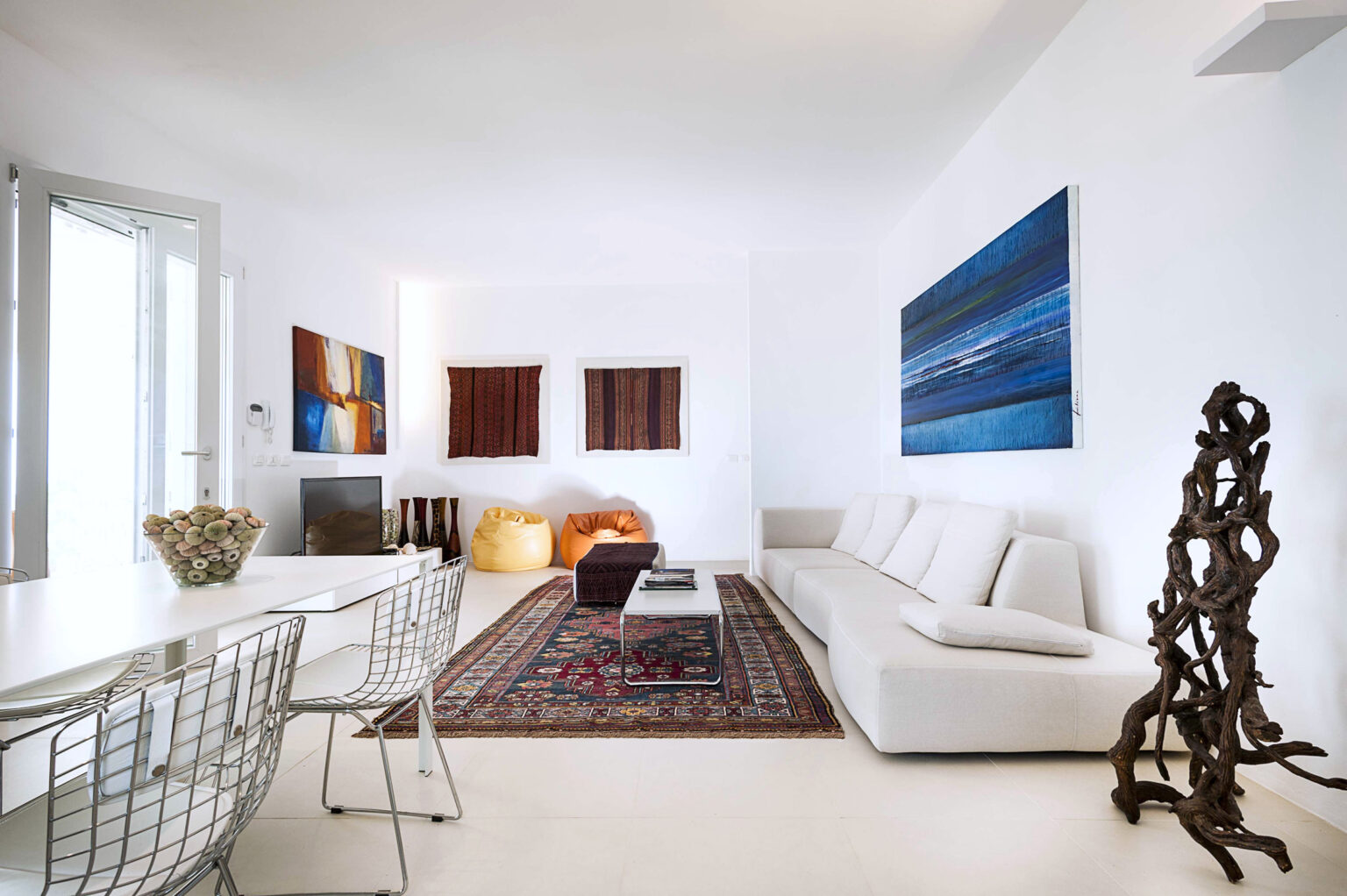 Modern living room featuring a white sofa, a decorative area rug, and contemporary art on the walls. The space includes a dining table with wire chairs and a cozy reading nook with a bean bag, all set in a bright, minimalist design. | Only on SopranoVillas