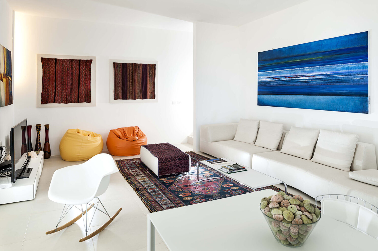 Modern living room featuring a white sectional sofa, colorful bean bags, a decorative rug, and a large blue abstract painting on the wall. The space is bright and airy, with minimalist decor and a stylish coffee table. | Only on SopranoVillas