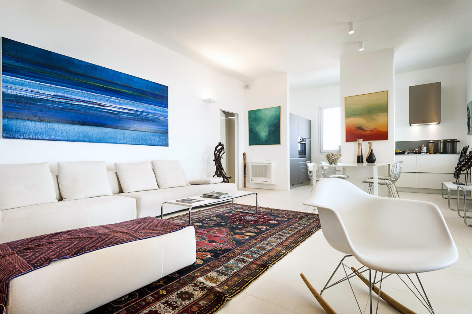 Modern living room featuring a white sectional sofa, colorful abstract paintings on the walls, a stylish coffee table, and a decorative area rug. The open layout includes a dining area and kitchen, emphasizing a bright and airy atmosphere. | Only on SopranoVillas
