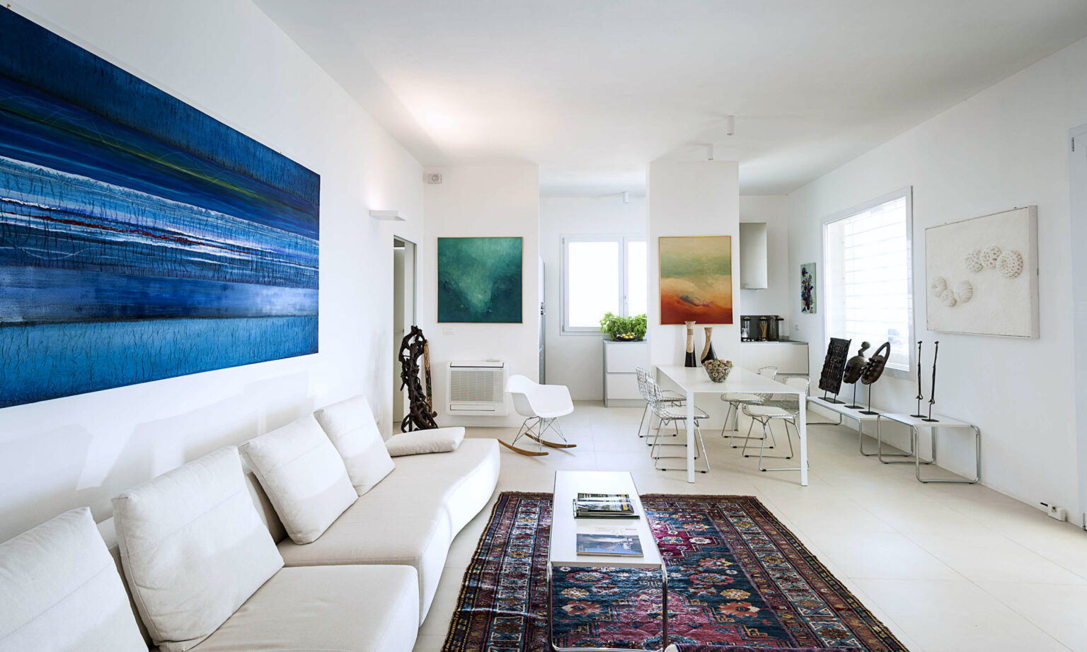 Modern living room featuring a white sofa, colorful abstract wall art, a decorative rug, and minimalist furniture. Natural light illuminates the space, enhancing the serene atmosphere. | Only on SopranoVillas