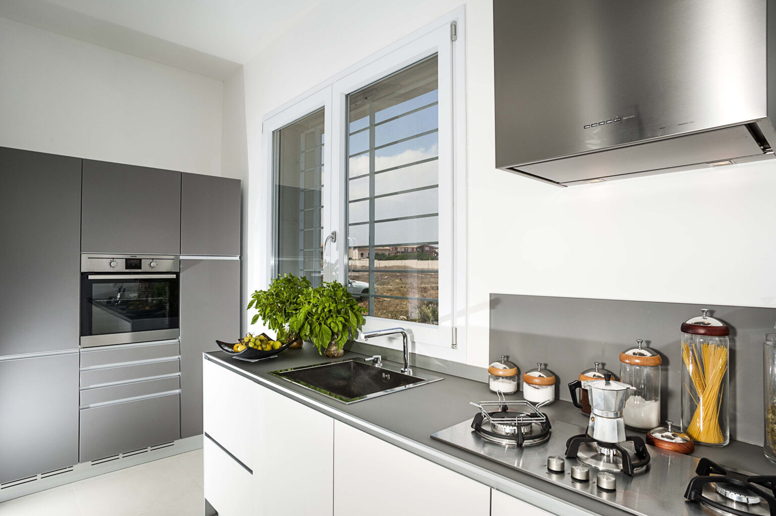 Modern kitchen featuring sleek gray cabinetry, stainless steel appliances, a gas stove, and a large window with natural light. Fresh herbs are displayed on the countertop, enhancing the contemporary design. | Only on SopranoVillas