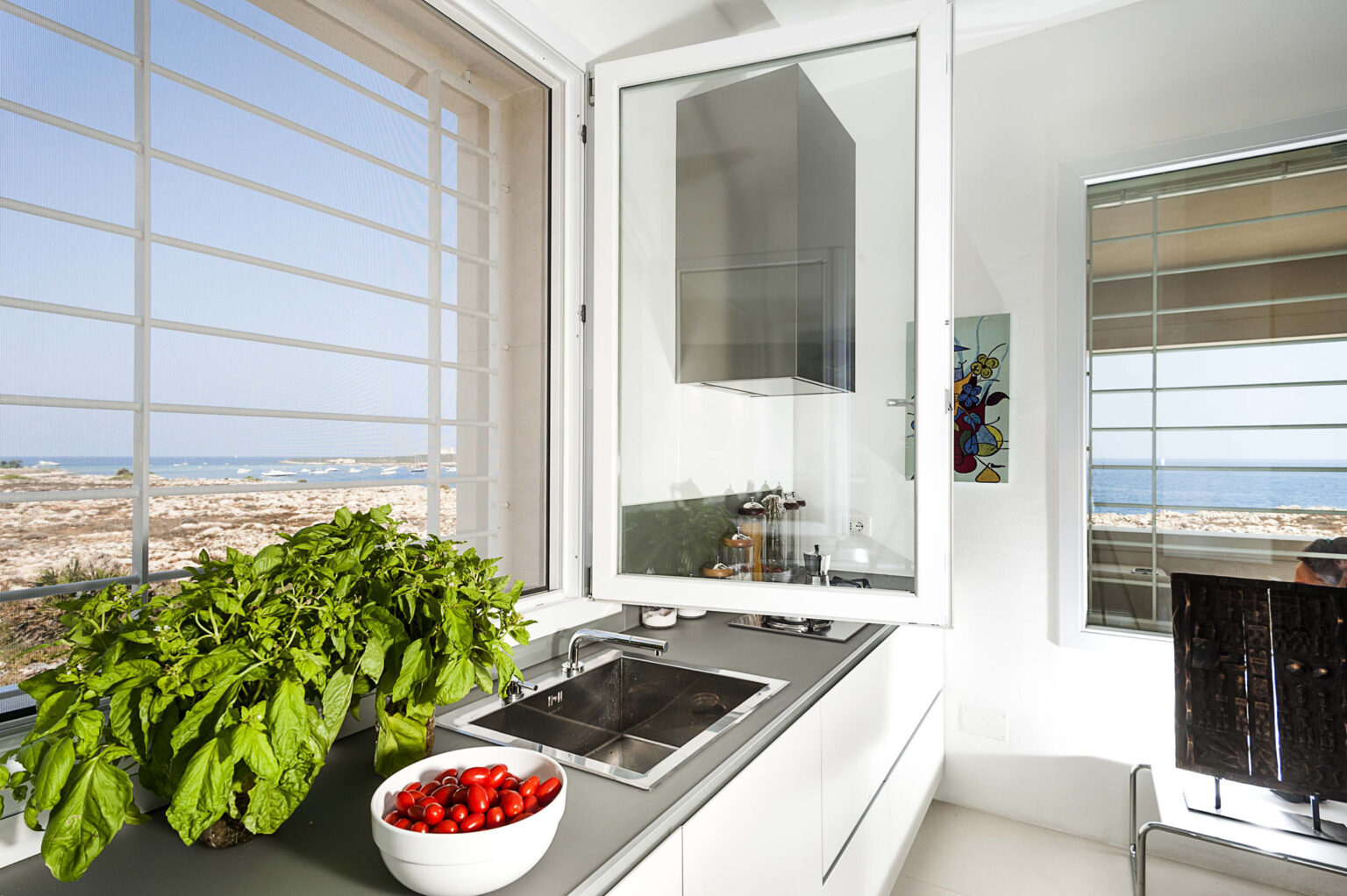 Modern kitchen with a scenic view, featuring a sleek countertop, built-in stove, fresh basil plant, and a bowl of cherry tomatoes. Natural light floods the space through large windows, enhancing the contemporary design. | Only on SopranoVillas